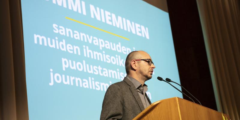 Artikkelin kuvituskuvassa haastateltava Tommi Nieminen pitämässä palkintopuhetta tiedonjulkistamisen valtionpalkintojen 2022 jakotilaisuudessa. Niemisen taustalla valkokankaalle heijastettuna teksti: "Tommi Nieminen. Sananvapauden ja muiden ihmisoikeuksien puolustamisesta journalismin keinoin."