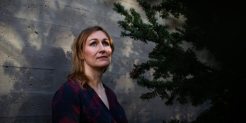 Kuvassa haastateltava Tiina Raevaara seisoo valkoista seinää vasten puun vieressä.