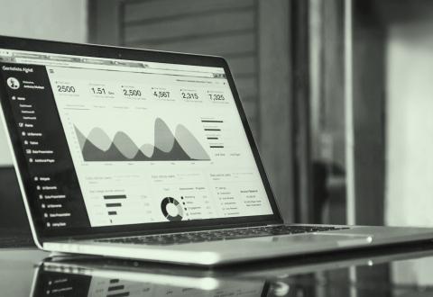 A laptop displaying an analytics and monitoring tool on the screen. Several charts and graphs are visible, including a line chart and a pie chart showing user data. The background features various shades of green.